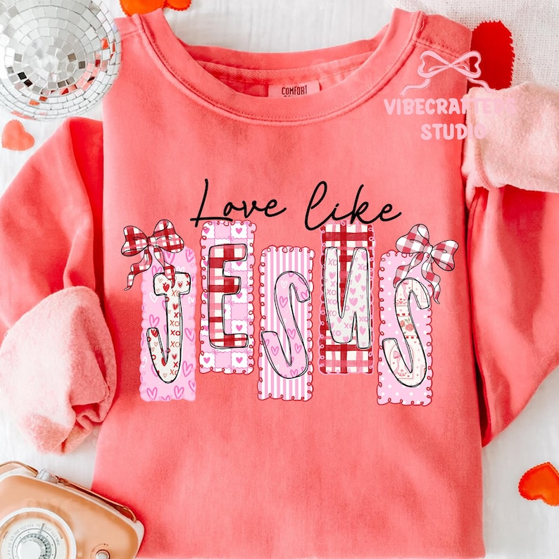 May include: A coral-colored sweatshirt with the phrase "Love like Jesus" in black script. The word "Jesus" is in a decorative, pink and red design with hearts and plaid patterns. A decorative bow is on each side of the word.
