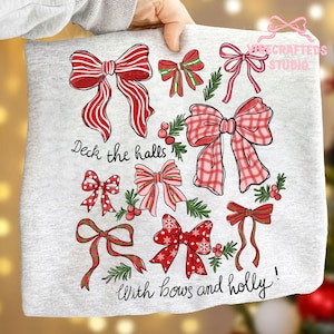 Może przedstawiać: Jasnoszara bluza z świątecznym wzorem różnych czerwonych kokardek z białymi akcentami i gałązkami ostrokrzewu. Tekst "Deck the halls" i "With bows and holly!" są również nadrukowane na bluzie, tworząc świąteczny motyw.