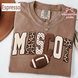 May include: An espresso-colored t-shirt featuring the word MASCOT in large, textured chenille letters with varying patterns including leopard print, polka dots, and solid brown. A brown football graphic is positioned below the text.