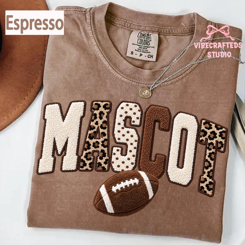 May include: An espresso-colored t-shirt featuring the word MASCOT in large, textured chenille letters with varying patterns including leopard print, polka dots, and solid brown. A brown football graphic is positioned below the text.