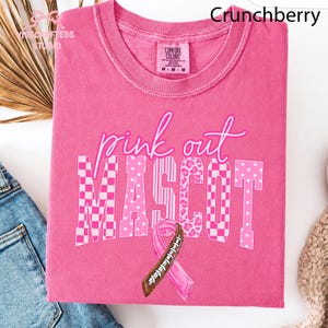 May include: A bright pink short-sleeve t-shirt featuring the text pink out MASCOT in white and pink patterns. The design includes a pink awareness ribbon combined with a brown and white football lace graphic. The shirt has a Comfort Colors label.