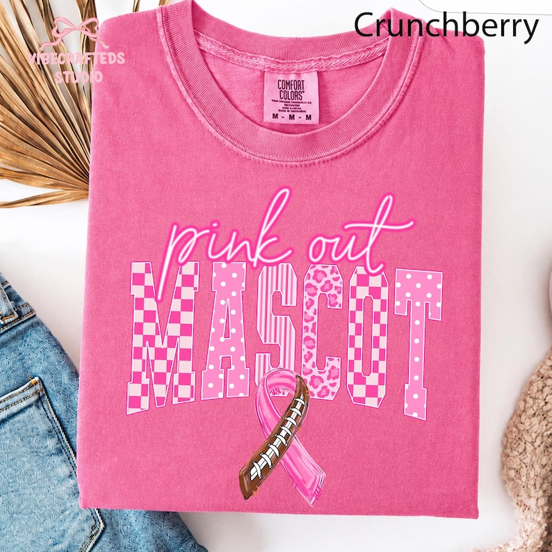 May include: A bright pink short-sleeve t-shirt featuring the text pink out MASCOT in white and pink patterns. The design includes a pink awareness ribbon combined with a brown and white football lace graphic. The shirt has a Comfort Colors label.