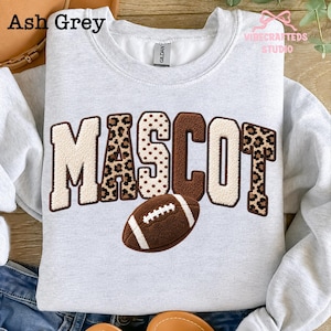 May include: An ash grey crewneck sweatshirt featuring the word MASCOT in large, textured varsity-style letters. The letters alternate between cream, brown, and leopard print patterns. A brown football graphic with white laces sits below the text.