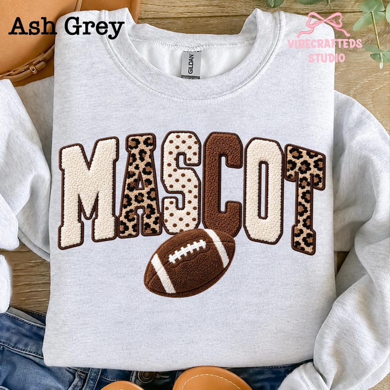 May include: An ash grey crewneck sweatshirt featuring the word MASCOT in large, textured varsity-style letters. The letters alternate between cream, brown, and leopard print patterns. A brown football graphic with white laces sits below the text.