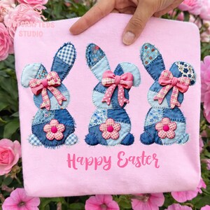 May include: A light pink Easter-themed shirt featuring three denim bunny appliqu&eacute;s with pink bows and flower accents. The bunnies have patchwork ears and bodies. The words "Happy Easter" are printed in pink.