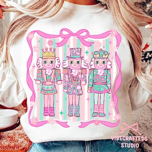 May include: White sweatshirt featuring a pink and green illustration of three nutcrackers in uniform. The design is framed by a pink ribbon with a bow at the top and the text "VIBECRAFTEDS STUDIO" at the bottom.