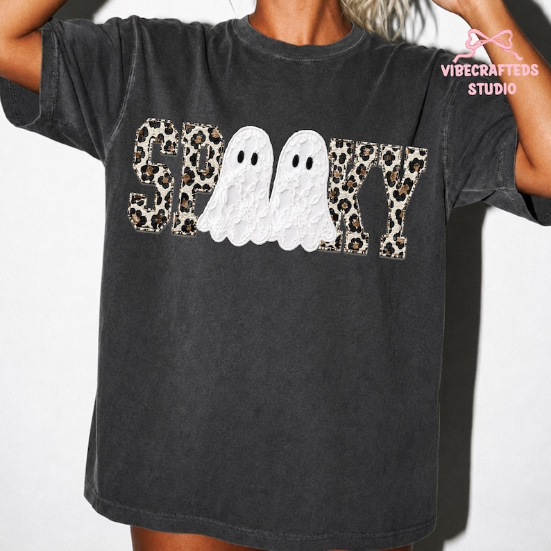 May include: A charcoal grey short-sleeve t-shirt featuring the text SPOOKY in a leopard print pattern. The two letter O's are replaced by white ghost illustrations with black eyes. The text VIBECRAFTEDS STUDIO appears in the top right corner.