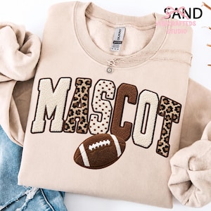 May include: A beige crewneck sweatshirt featuring the word MASCOT in large, textured chenille letters with varying patterns including leopard print, polka dots, and solid brown. A brown football with white laces is embroidered below the text.