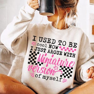 以下が含まれることがあります： 「I USED TO BE cool NOW I JUST ARGUE WITH Miniature version of myself」のテキストが黒とピンクでプリントされたクリーム色のスウェットシャツ。デザインには星、ハート、市松模様が含まれています。