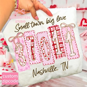 Könnte beinhalten: Weißes T-Shirt mit dem Text "Small town, big love" und "Nashville, TN". Das Design zeigt die Postleitzahl "37011" in rosa und roten Mustern mit goldenen Akzenten und Schleifen. Das T-Shirt wird von einer Hand gehalten.