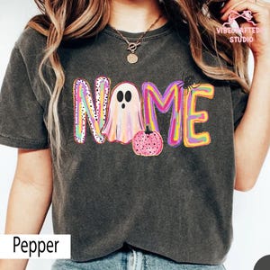 May include: A charcoal gray t-shirt featuring a colorful graphic with the word NAME, where the letter A is replaced by a ghost and a small pink pumpkin. The design includes vibrant swirls of yellow, pink, and purple, with a black spider accent.