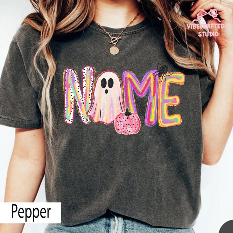 May include: A charcoal gray t-shirt featuring a colorful graphic with the word NAME, where the letter A is replaced by a ghost and a small pink pumpkin. The design includes vibrant swirls of yellow, pink, and purple, with a black spider accent.