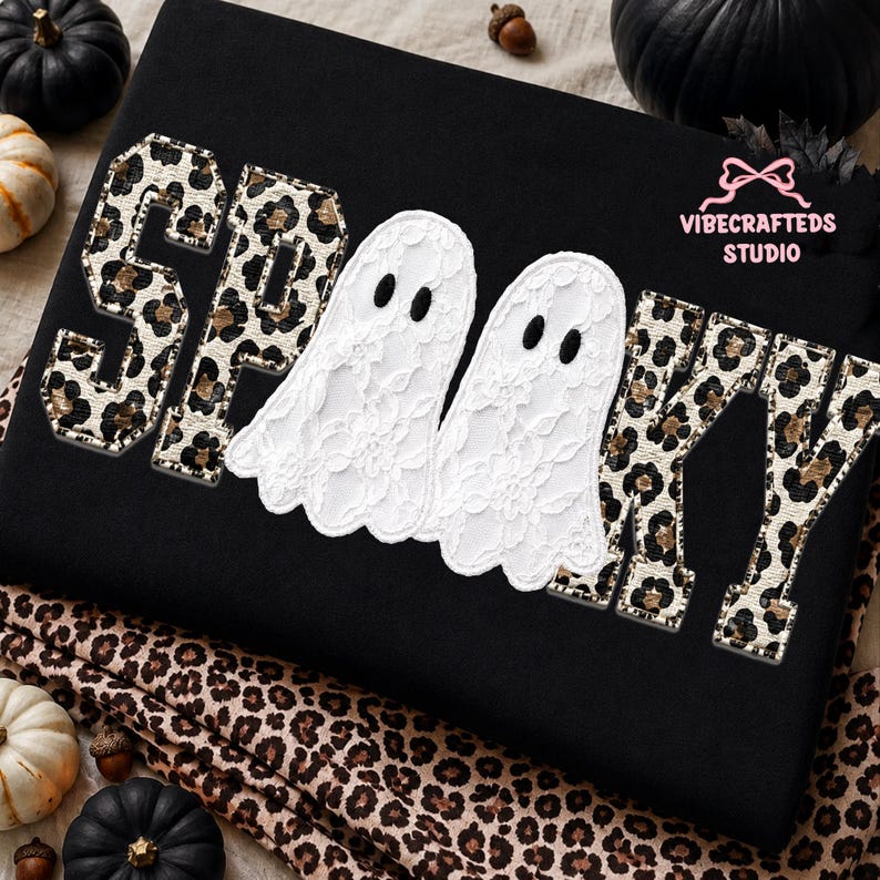 May include: A black sweatshirt featuring the word SPOOKY in varsity-style letters with a leopard print pattern. The two O's are replaced by white lace ghost appliqu&eacute;s with black oval eyes. The text VIBECRAFTEDS STUDIO appears in pink.