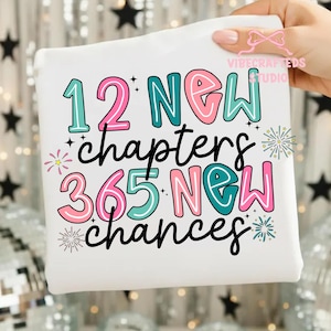 May include: White sweatshirt with the text "12 NEW chapters 365 NEW chances" in teal, pink, and black. The design includes star and firework graphics. The sweatshirt is held up by a person.