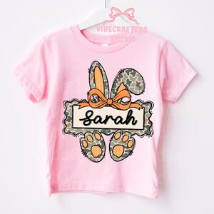 May include: A pink t-shirt featuring a bunny design with the name "Sarah". The bunny ears and paws have a camouflage pattern with orange accents. The name is written in a cursive font.