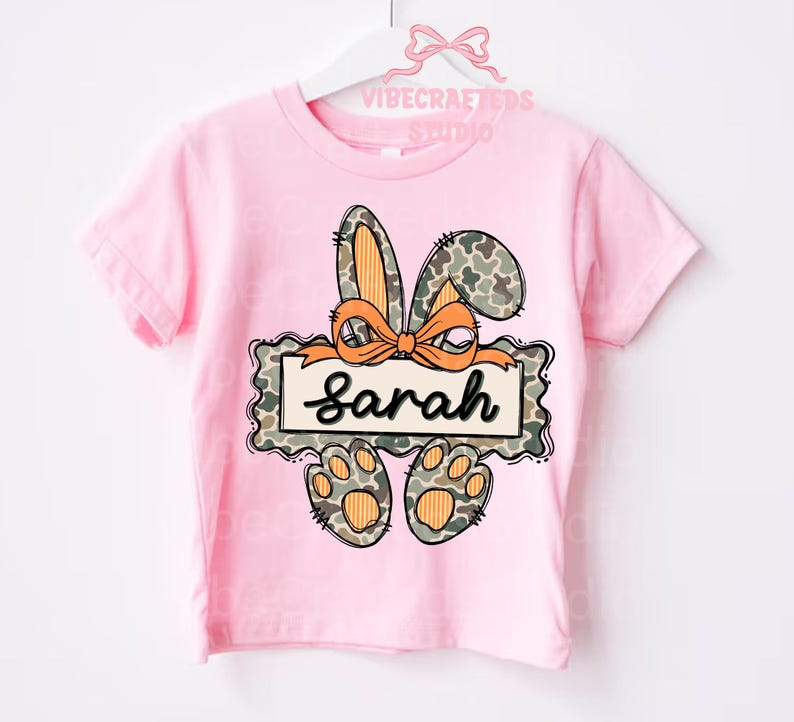 May include: A pink t-shirt featuring a bunny design with the name "Sarah". The bunny ears and paws have a camouflage pattern with orange accents. The name is written in a cursive font.