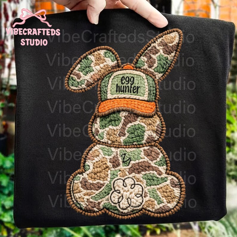 May include: Black fabric with an embroidered bunny design. The bunny wears a camouflage hat with the text "egg hunter". The bunny's body is also in camouflage. A four-leaf clover is at the bottom.