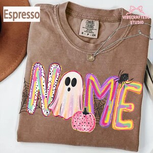May include: An espresso-colored t-shirt featuring a graphic with the word NAME, where the letter A is replaced by a ghost, accompanied by a pink pumpkin, a black spider, and spiderweb patterns in vibrant, multi-colored, paint-stroke style.
