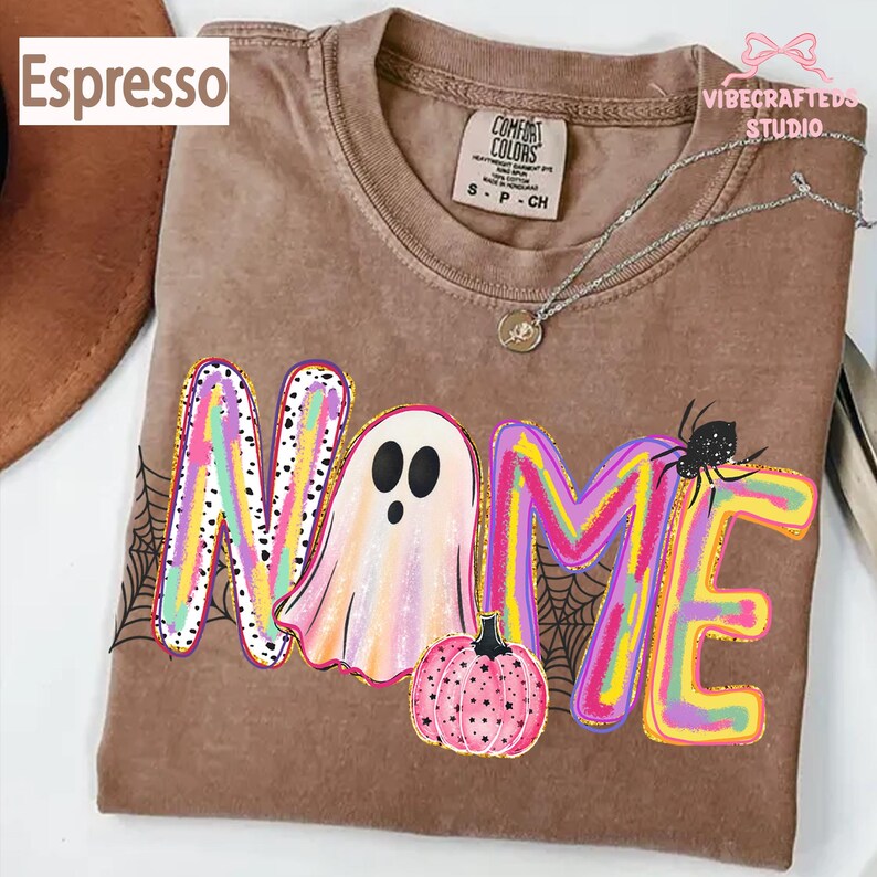 May include: An espresso-colored t-shirt featuring a graphic with the word NAME, where the letter A is replaced by a ghost, accompanied by a pink pumpkin, a black spider, and spiderweb patterns in vibrant, multi-colored, paint-stroke style.