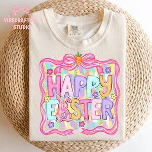 May include: Cream-colored t-shirt with a colorful "Happy Easter" graphic. The design features a pink bunny, flowers, and a bow with a carrot. The text is in various pastel colors, framed by a pink border with gold glitter accents.