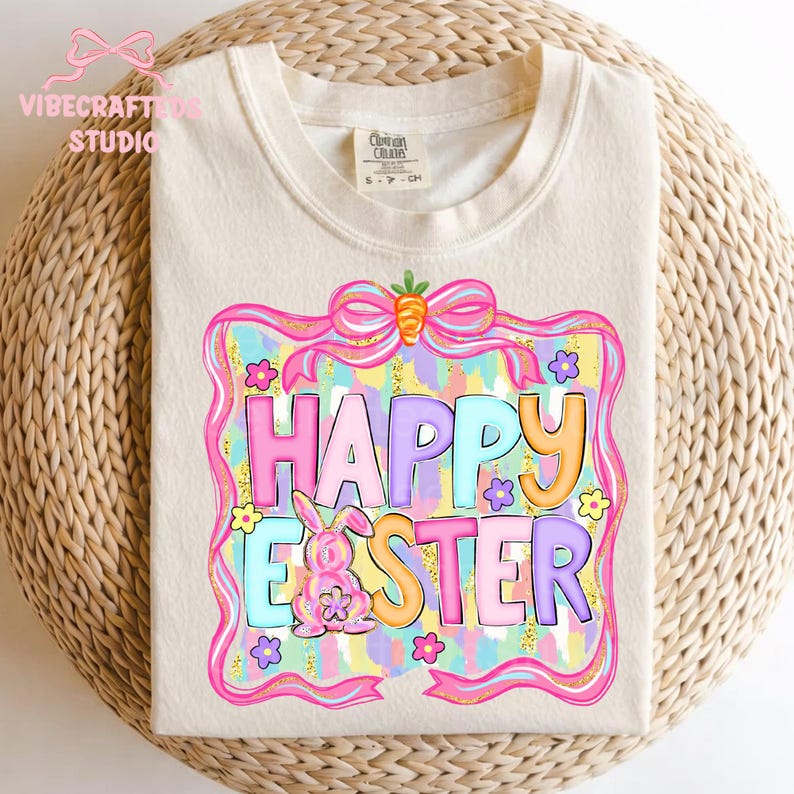 May include: Cream-colored t-shirt with a colorful "Happy Easter" graphic. The design features a pink bunny, flowers, and a bow with a carrot. The text is in various pastel colors, framed by a pink border with gold glitter accents.