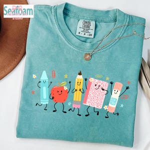 Puede incluir: Una camiseta de manga corta color verde azulado con un estampado gráfico de cinco útiles escolares antropomórficos, incluyendo un crayón azul, una manzana roja, un lápiz amarillo, un cuaderno rosa y un pegamento, todos con caras sonrientes.
