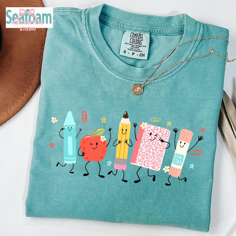 Puede incluir: Una camiseta de manga corta color verde azulado con un estampado gr&aacute;fico de cinco &uacute;tiles escolares antropom&oacute;rficos, incluyendo un cray&oacute;n azul, una manzana roja, un l&aacute;piz amarillo, un cuaderno rosa y un pegamento, todos con caras sonrientes.