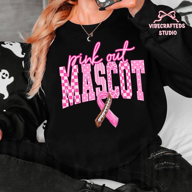 May include: A black long-sleeve sweatshirt featuring the pink text "pink out" above the word "MASCOT" in a large, blocky font with various pink patterns including checkers, polka dots, and leopard print, plus a pink awareness ribbon football graphic.