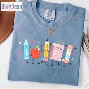 Puede incluir: Una camiseta azul de cuello redondo con un estampado gr&aacute;fico de cinco &uacute;tiles escolares antropom&oacute;rficos, incluyendo un cray&oacute;n azul claro, una manzana roja, un l&aacute;piz amarillo, un cuaderno rosa y un pegamento en barra, todos con caras sonrientes.