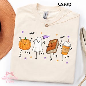 May include: A sand-coloured t-shirt featuring a graphic print of a smiling pumpkin, a ghost holding a purple pennant that says SPOOKY, a brown book labelled NOVEL, and an orange iced coffee cup, all surrounded by small purple and orange stars and bats.