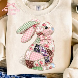 May include: Cream-colored sweatshirt featuring a patchwork bunny design. The bunny is made of various patterned fabrics, including gingham, floral, and plaid. It has a pink bow and a fluffy white tail. The sweatshirt has the text "Vibe Crafted Studio".