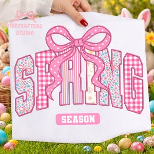Könnte beinhalten: Weißer Stoff mit dem Wort "SPRING" im College-Stil, mit Blumen-, Streifen- und Vichy-Mustern. Über dem Wort befindet sich eine große rosa Schleife mit Punkten. Das Wort "SEASON" steht darunter. Das Bild wird von einer Person gehalten.
