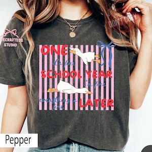 May include: Dark gray t-shirt with a graphic featuring two white ducks and the text "ONE SCHOOL YEAR I'M FINE EVERYTHING IS LATER". The design includes pink and white stripes and a blue ribbon.