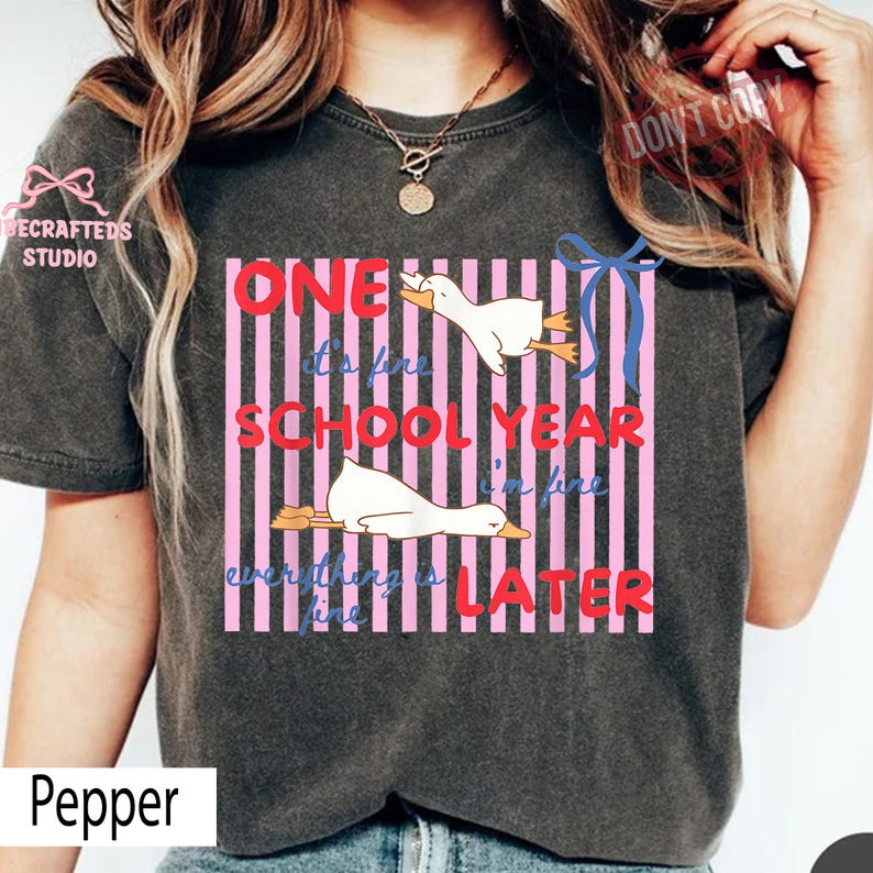 May include: Dark gray t-shirt with a graphic featuring two white ducks and the text "ONE SCHOOL YEAR I'M FINE EVERYTHING IS LATER". The design includes pink and white stripes and a blue ribbon.