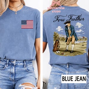 May include: A blue t-shirt featuring a small American flag on the front chest. The back displays a graphic of a historical figure in colonial attire playing golf with the text Fore Father above, and a golf bag with clubs on the green grass.