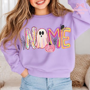 May include: A light purple crewneck sweatshirt featuring a colorful graphic with the word NAME, where the letter A is replaced by a ghost, a pink polka-dot pumpkin, black spiderwebs, and a small black spider. The text BECRAFTEDS STUDIO is visible.