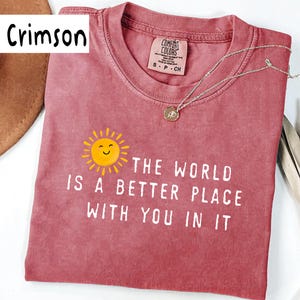 May include: A crimson short-sleeve t-shirt featuring a graphic of a smiling yellow sun and the white text "THE WORLD IS A BETTER PLACE WITH YOU IN IT". The shirt has a crew neckline and a Comfort Colors brand label inside the collar showing size S.