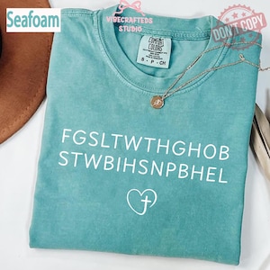 Könnte beinhalten: Meergrünes T-Shirt mit weißem Text und einer Herz-Kreuz-Grafik. Das Shirt hat einen Rundhalsausschnitt und kurze Ärmel. Eine goldene Halskette mit Anhänger liegt auf dem Shirt. Das Shirt ist mit "Comfort Colors" gekennzeichnet.