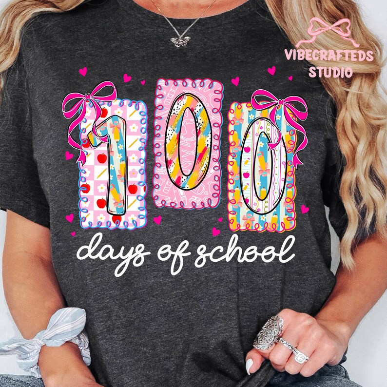 May include: Dark gray t-shirt with a colorful graphic celebrating 100 days of school. The design features the numbers "100" in a playful font, decorated with bows, hearts, and school-themed illustrations. The text "days of school" is written below.