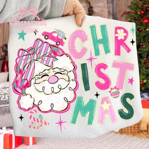 May include: White sweatshirt with a colorful Christmas design. The design features a Santa Claus with a pink and green hat and bow, a pink car with a Christmas tree, and the word "CHRISTMAS" in pink and green letters.