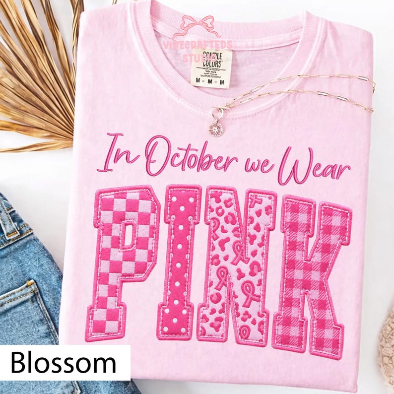 May include: A light pink t-shirt featuring the text In October we Wear in dark pink script above the word PINK in large block letters. Each letter of PINK displays a different pattern including checkers, polka dots, leopard print, and plaid.