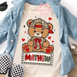 May include: A tan t-shirt featuring a cartoon teddy bear wearing a camouflage baseball cap and holding a heart-shaped gift with a red bow. The name "MATHEW" is printed on a sign below the bear. Red hearts and black stars are scattered around the bear.