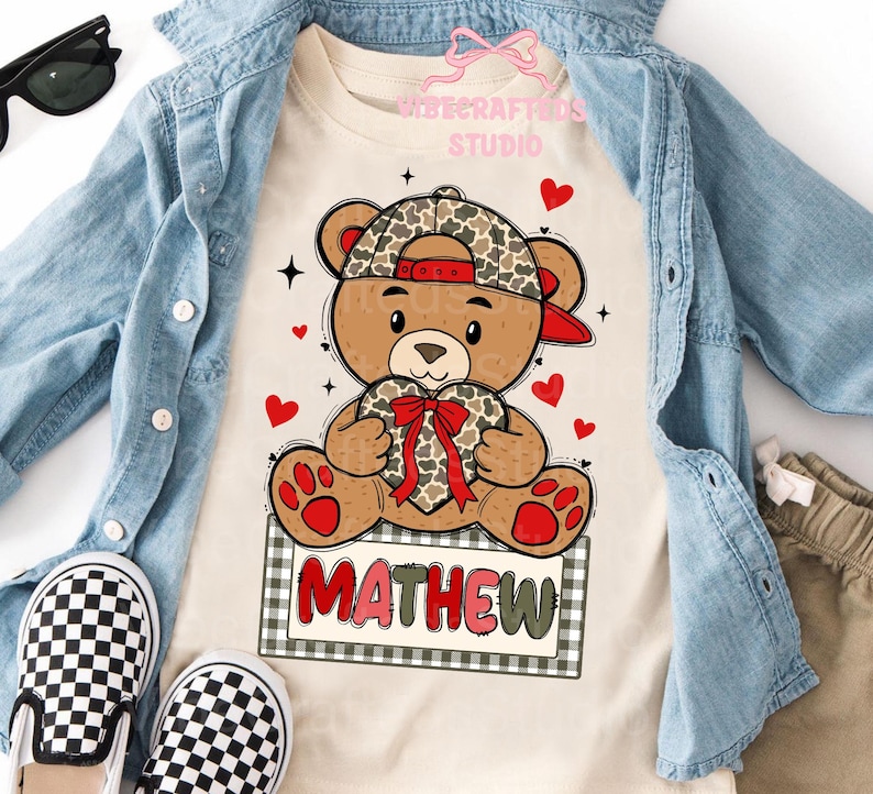 May include: A tan t-shirt featuring a cartoon teddy bear wearing a camouflage baseball cap and holding a heart-shaped gift with a red bow. The name "MATHEW" is printed on a sign below the bear. Red hearts and black stars are scattered around the bear.