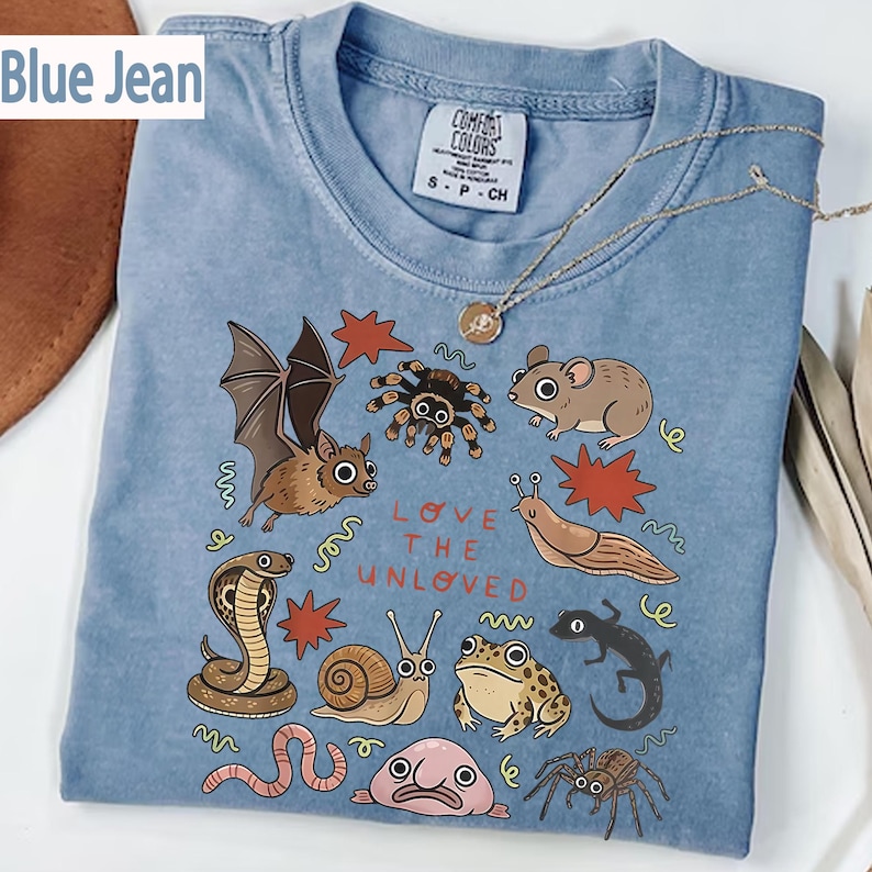 May include: A blue t-shirt featuring a colourful graphic of creatures such as a bat, spider, and mouse. The text "LOVE THE UNLOVED" is printed on the shirt. A gold necklace is draped over the shirt.