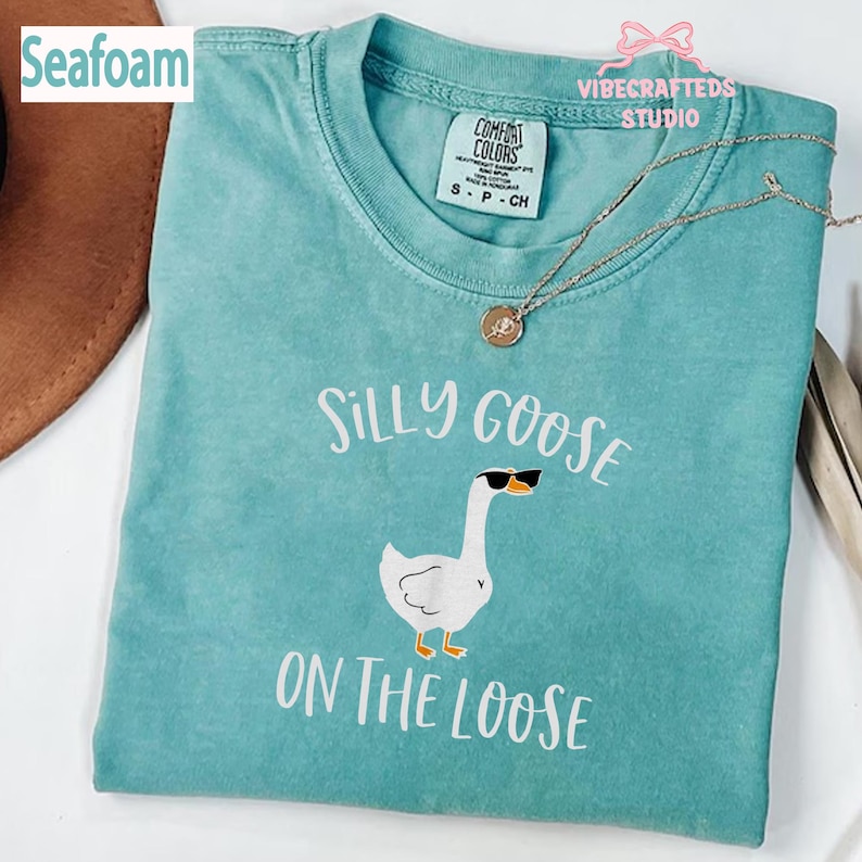 May include: Seafoam green t-shirt with the phrase "Silly Goose On The Loose" and a cartoon goose wearing sunglasses. The shirt has a crew neck and short sleeves. A silver necklace with a pendant is draped on the shirt.