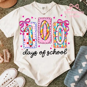 May include: A white t-shirt with a colorful graphic celebrating 100 days of school. The design features the numbers "100" in a playful font, decorated with bows, hearts, and school-themed elements. The words "days of school" are written in a cursive font.
