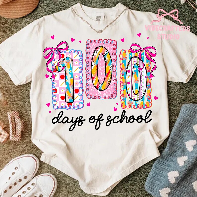 May include: A white t-shirt with a colorful graphic celebrating 100 days of school. The design features the numbers "100" in a playful font, decorated with bows, hearts, and school-themed elements. The words "days of school" are written in a cursive font.