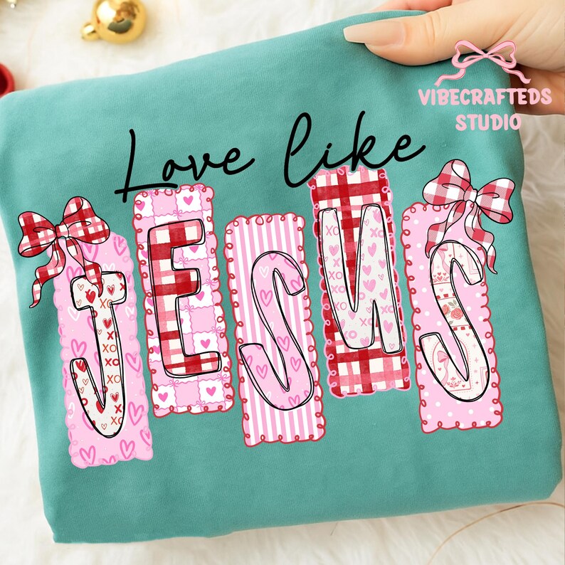 May include: Teal sweatshirt with the words "Love like JESUS" in black script. The word JESUS is in pink and red patterned letters with bows and hearts. A hand is holding the sweatshirt.