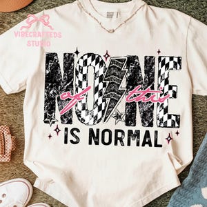 May include: Cream-coloured t-shirt featuring the text "NONE of this IS NORMAL" in a distressed black and white chequered design. A pink lightning bolt and text accent the design. The shirt has a V-neck.