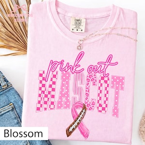 May include: A light pink short-sleeved t-shirt featuring the text pink out MASCOT in various pink patterns including checkerboard, stripes, leopard print, and polka dots. A pink awareness ribbon designed with football laces is centered below the text.